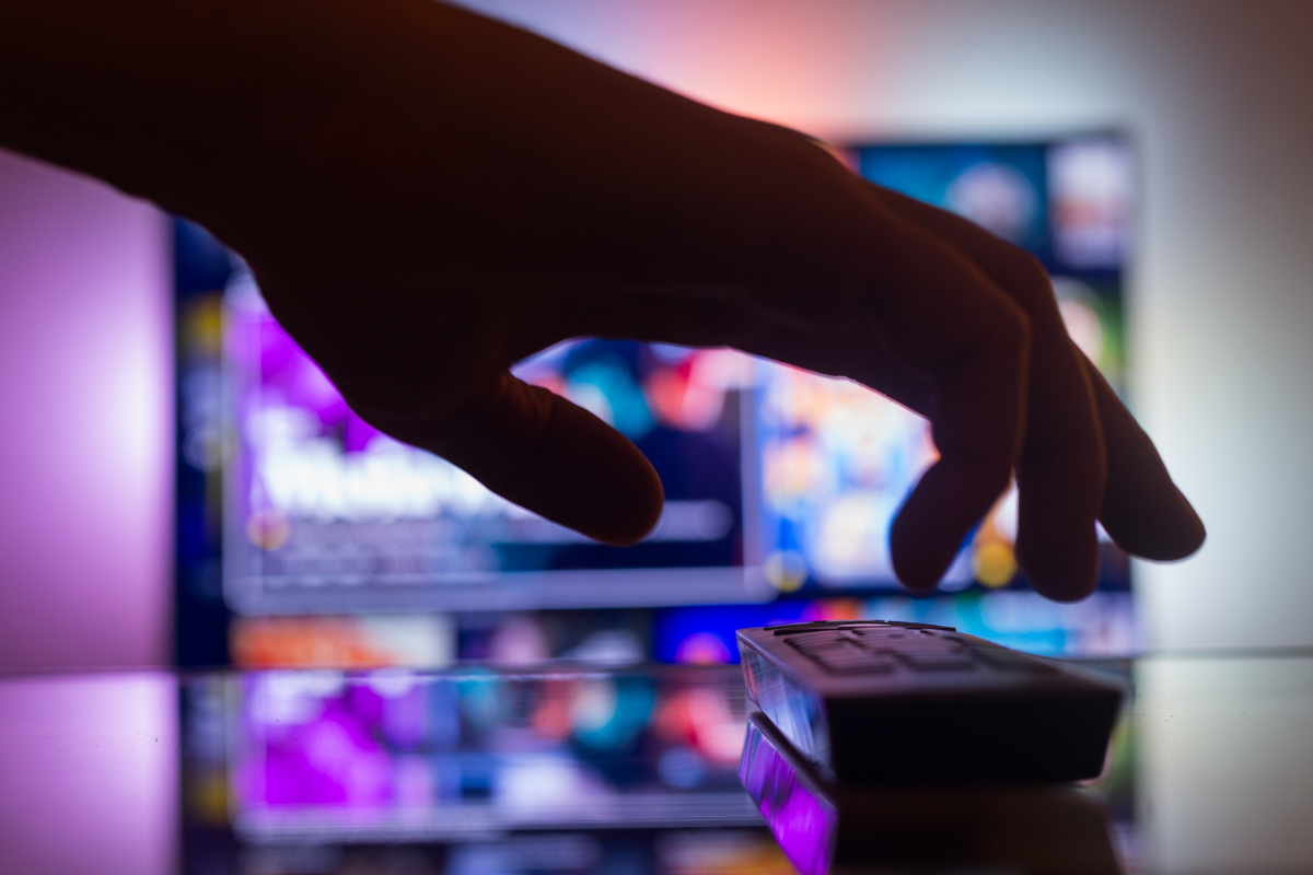 A close-up, stylized view of a hand reaching for a television remote in front of a glowing smart TV streaming interface, symbolizing the highly targeted CTV ad environment for businesses.
